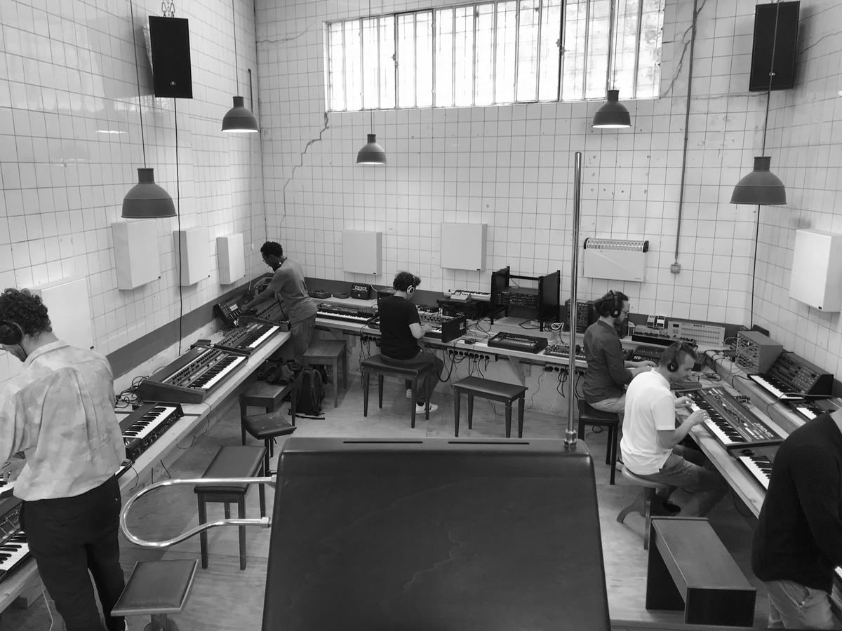 Schwarz-Weiss Foto eines gekachelten Raumes. Sieben Menschen beugen sich über verschiedene elektronische Musikinstrumente.